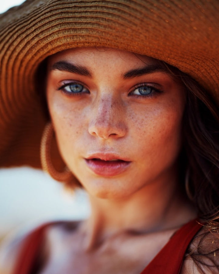Kvindeansigt med solhat, der skal illustrerer en der bruger hudplejeroduktet Ainhoa Skin Primers SPF50: Let og fugtgivende ansigtsemulsion med anti-age effekt. Reducerer rynker og beskytter mod blåt lys og forurening.