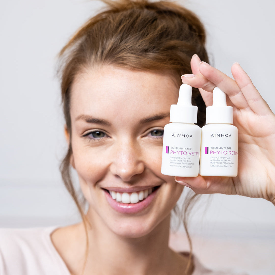 Billedet viser et kvindeansigt der holder to hudplejeprodukter i hånden. Hudplejeprodukterne er Ainhoa Phyto Retin+ Facial Oil: Klinisk testet ansigtsolie for rynker og elasticitet. Naturlig og vegansk pleje til tør hud. Anvendes alene eller med creme.